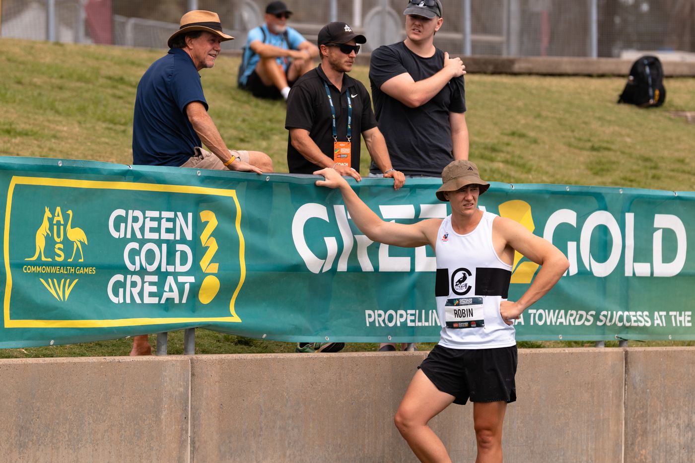 An athlete in a club singlet talking to coaches leaning over the trackside barrier at a meet.