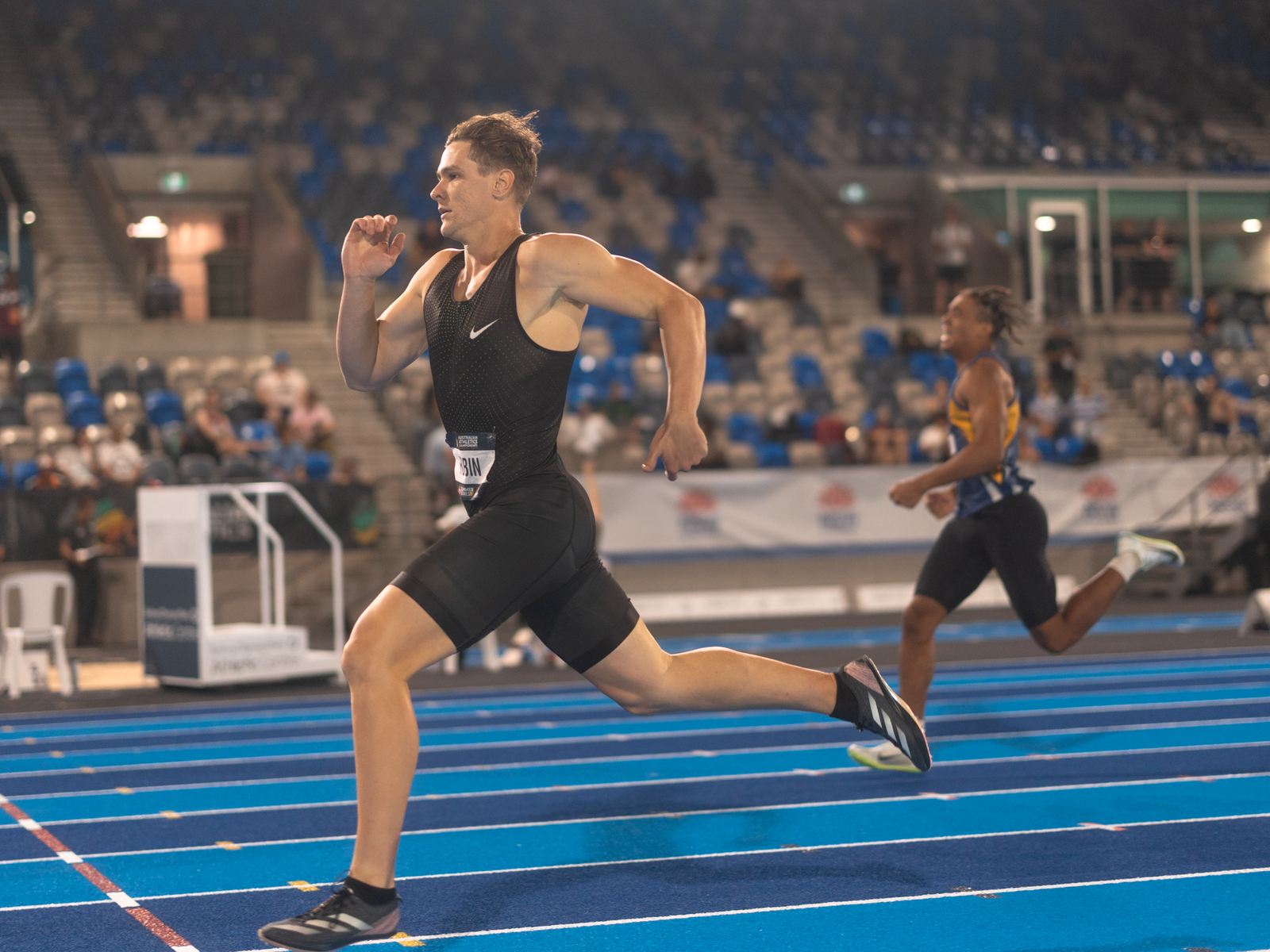 A sprinter at full stride down the home straight under stadium lights, a second athlete behind him.